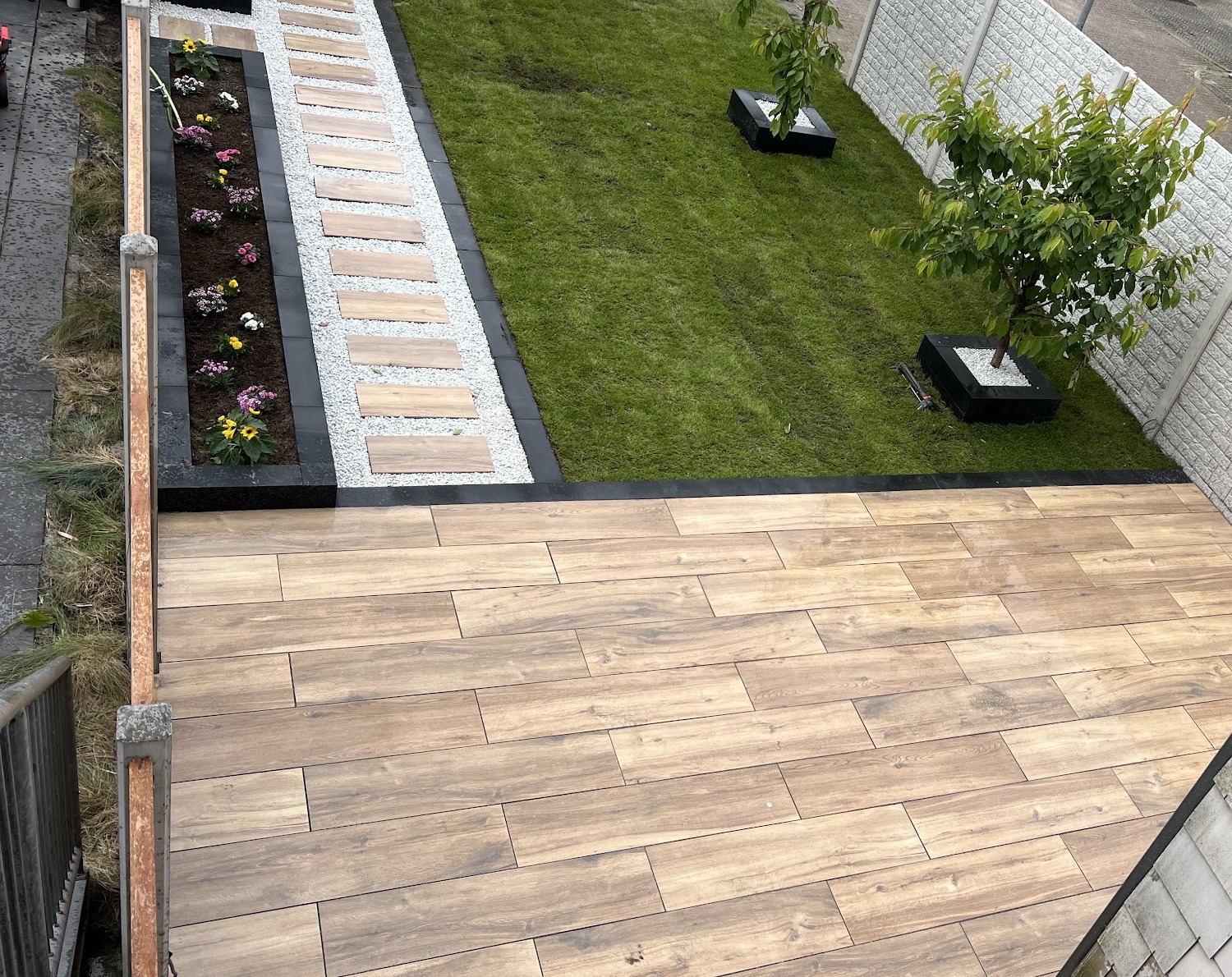 Terras in houtlook keramische tegels, gazon, een stapstenenpad in wit grind en een plantenbed