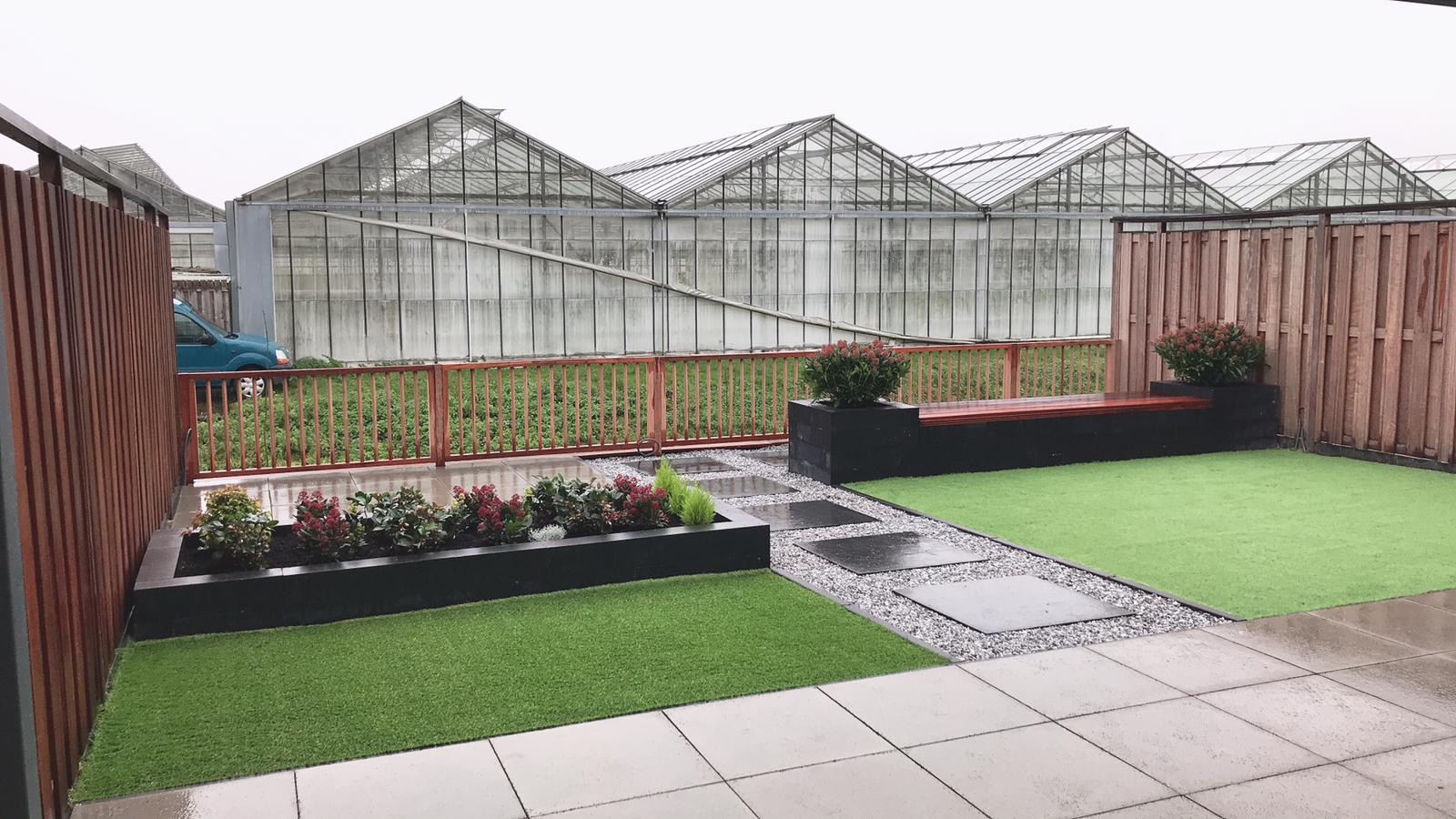 Tuin met kunstgras, zwarte plantenbakken en stapstenen in grind, met kassen op de achtergrond