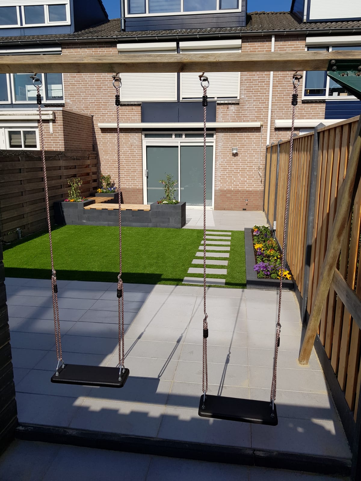 Terras met grijze tegels, twee schommels aan een houten balk, een klein gazon met stapstenen en een bloemenborder