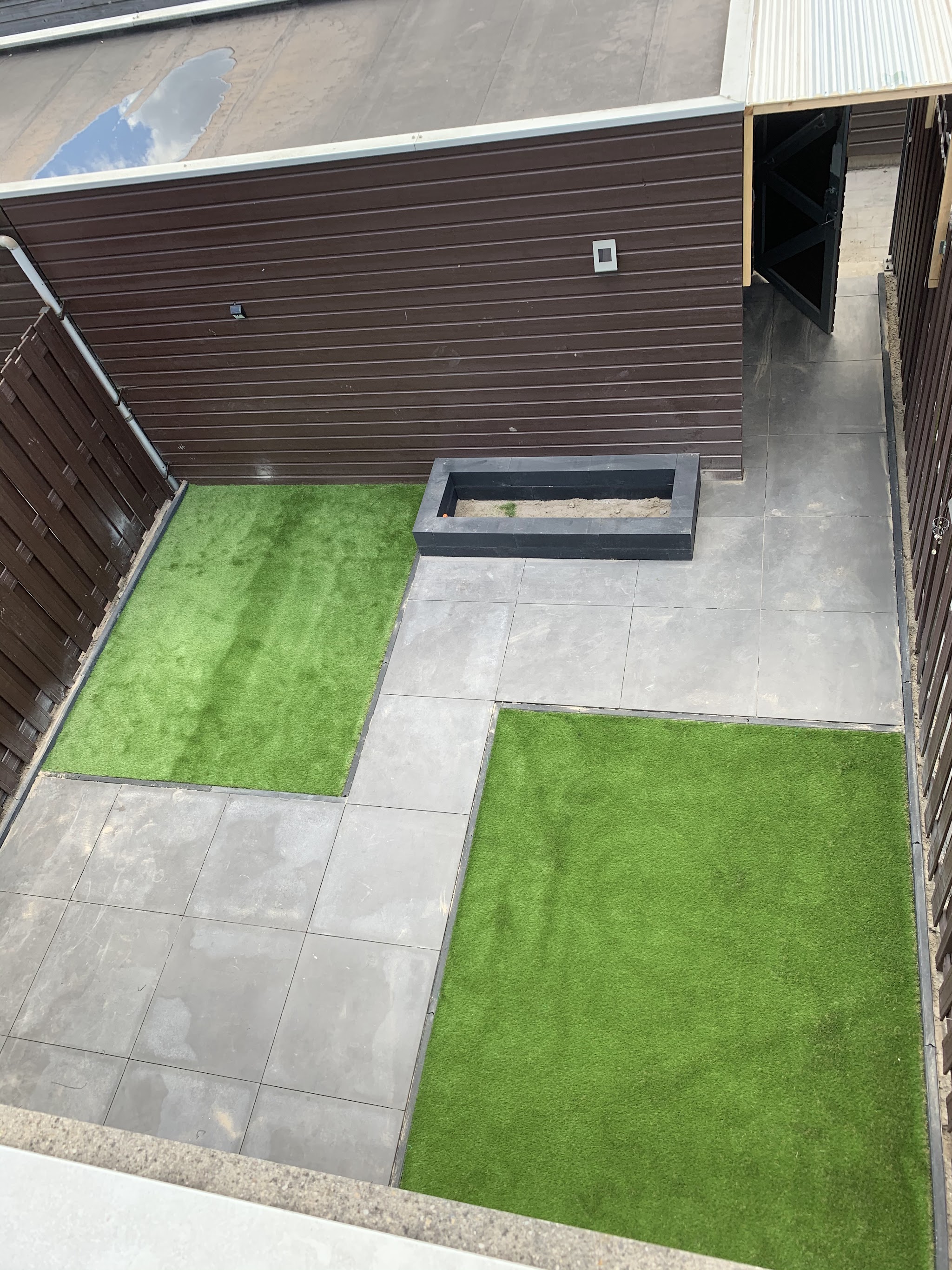 Kleine tuin met vlakken kunstgras, grijze tegels en een zandbak, van boven gezien